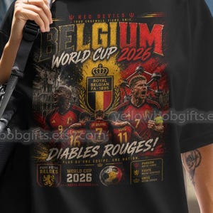 May include: A black short-sleeved t-shirt featuring a graphic print with the text Belgium World Cup 2026 and Diables Rouges. The design includes illustrations of football players, the Royal Belgian FA crest, and various tournament-related details.