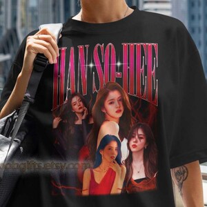 Könnte beinhalten: Schwarzes T-Shirt mit einer Grafik von Han So Hee und dem Text "HAN SO HEE" in einer Retro-Schriftart. Das T-Shirt hat einen Rundhalsausschnitt und kurze Ärmel. Das Bild ist vor dem Hintergrund einer Stadtlandschaft platziert.
