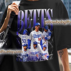 May include: Black t-shirt featuring a graphic of baseball players in blue and white uniforms. The shirt has the word "BETTS" in large, purple letters across the top. The text "Dodgers" is also visible. The Etsy shop name "yellowbobgifts.etsy.com" is printed on the shirt.
