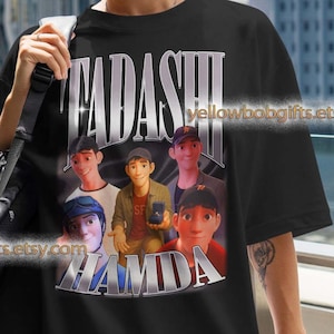 Puede incluir: Camiseta negra con un gráfico de personajes animados y el texto "FADASHI HAMDA". El diseño incluye imágenes de varias personas. La camiseta la lleva una persona con una bolsa negra.