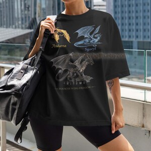 K&ouml;nnte beinhalten: Schwarzes T-Shirt mit drei Drachenillustrationen, beschriftet mit Andarna, Sgaeyl und einem dritten Drachen. Der Text 'Fourth Wing Dragon' befindet sich unten. Das Shirt wird mit schwarzen Shorts und einer schwarzen Umh&auml;ngetasche getragen.