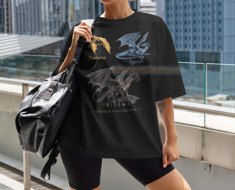 K&ouml;nnte beinhalten: Schwarzes T-Shirt mit drei Drachenillustrationen, beschriftet mit Andarna, Sgaeyl und einem dritten Drachen. Der Text 'Fourth Wing Dragon' befindet sich unten. Das Shirt wird mit schwarzen Shorts und einer schwarzen Umh&auml;ngetasche getragen.