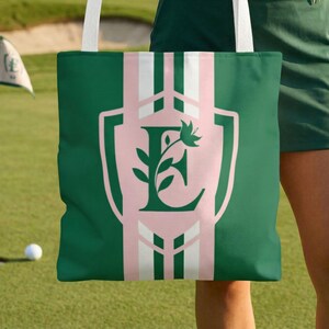 May include: Green tote bag with a pink and white striped design. The bag features a shield with the letter "E" and a floral design. The bag has white straps and is held on a golf course.