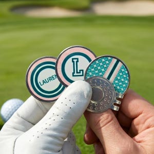 Könnte beinhalten: Eine Sammlung personalisierter Golfballmarker. Die Marker weisen eine Vielzahl von Designs auf, darunter ein türkis-rosa Farbschema, der Buchstabe "L" und der Name "Lauren". Ein Golfball und grünes Gras sind im Hintergrund.