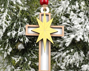 May include: Wooden Christmas ornament in the shape of a cross with a yellow starburst design. The cross has a light wood frame with a silver center. A red bead and a white ring are attached to the top with a natural twine loop for hanging.