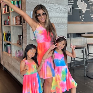 May include: A woman and two girls wearing tie-dye dresses. The woman is wearing a pink and blue tie-dye dress, and the girls are wearing pink and blue tie-dye dresses. They are all smiling and looking at the camera. The woman is holding her hand up in the air, and the girls are holding their hands up in the air. The background is a coffee shop with a chalkboard wall.