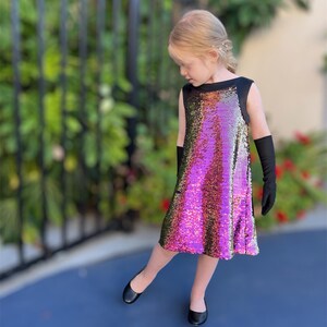 May include: A young girl wearing a sleeveless dress with a black bodice and a sequined skirt in shades of pink, gold, and green. She is wearing black gloves and black flats.