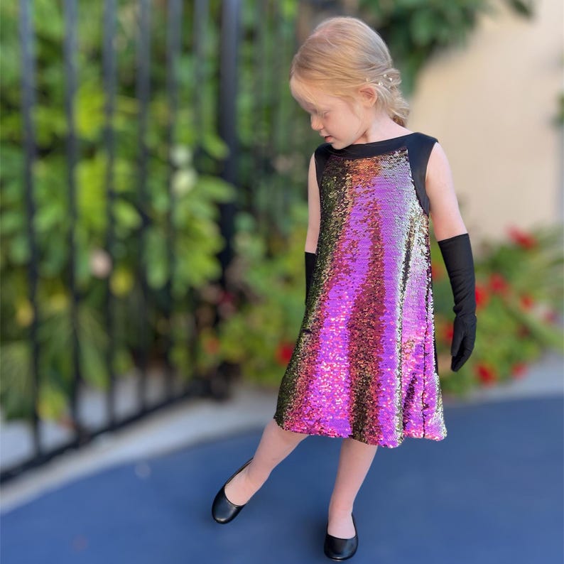 May include: A young girl wearing a sleeveless dress with a black bodice and a sequined skirt in shades of pink, gold, and green. She is wearing black gloves and black flats.