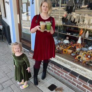 May include: A young girl in a green velvet dress stands next to a woman in a red velvet dress holding three takeaway cups of coffee and smoothies. The woman is standing in front of a storefront with the words "crystals jewelry art" and "thank you" visible in the window.