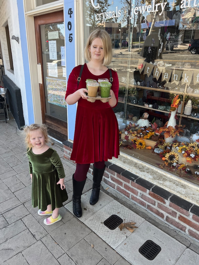 May include: A young girl in a green velvet dress stands next to a woman in a red velvet dress holding three takeaway cups of coffee and smoothies. The woman is standing in front of a storefront with the words "crystals jewelry art" and "thank you" visible in the window.