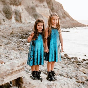 May include: Two girls wearing blue and black sequined dresses and black boots stand on a rock by the ocean.