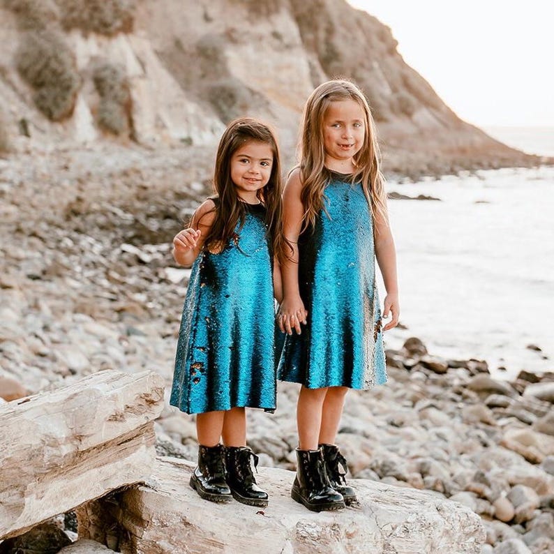 May include: Two girls wearing blue and black sequined dresses and black boots stand on a rock by the ocean.