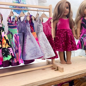 May include: A wooden clothes rack with a selection of colourful dresses hanging on it. Two dolls are standing in front of the rack, each wearing a pink dress. The doll on the left is wearing a pink velvet dress with sequins, and the doll on the right is wearing a pink and black patterned dress.