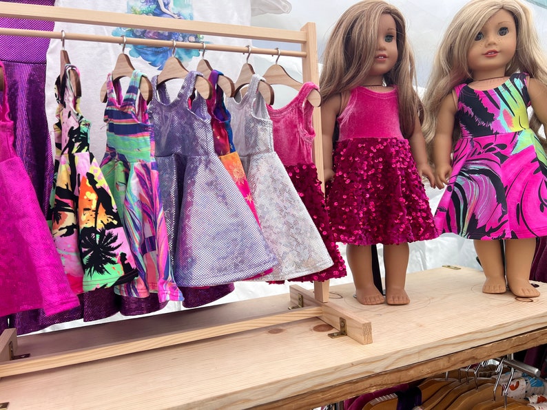 May include: A wooden clothes rack with a selection of colourful dresses hanging on it. Two dolls are standing in front of the rack, each wearing a pink dress. The doll on the left is wearing a pink velvet dress with sequins, and the doll on the right is wearing a pink and black patterned dress.