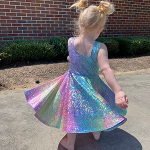 May include: A young girl wearing a rainbow iridescent dress with a twirling skirt. The dress has a sleeveless design and a fitted bodice. The girl has her hair pulled back in two pigtails.