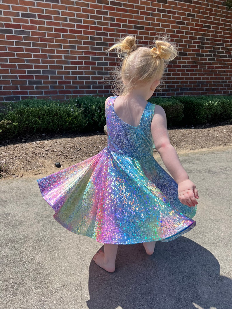 May include: A young girl wearing a rainbow iridescent dress with a twirling skirt. The dress has a sleeveless design and a fitted bodice. The girl has her hair pulled back in two pigtails.