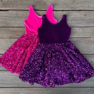May include: Two layered girls' dresses with sequined skirts. The top layer of the dress on the left is pink velvet, and the top layer of the dress on the right is purple velvet.