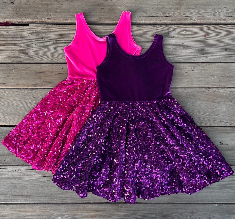 May include: Two layered girls' dresses with sequined skirts. The top layer of the dress on the left is pink velvet, and the top layer of the dress on the right is purple velvet.