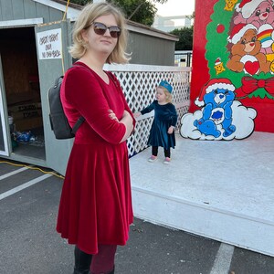 May include: A woman wearing a red velvet dress and black boots stands in front of a painted backdrop featuring a blue Care Bear with a white cloud. A young girl in a blue dress stands in front of the woman.