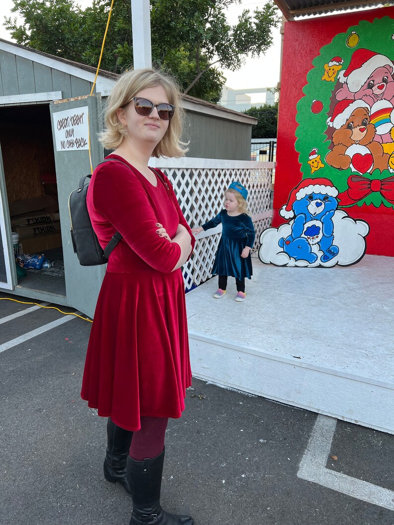 May include: A woman wearing a red velvet dress and black boots stands in front of a painted backdrop featuring a blue Care Bear with a white cloud. A young girl in a blue dress stands in front of the woman.