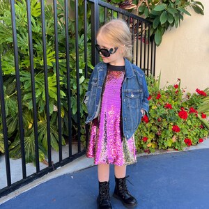 May include: A young girl wearing a pink and gold sequined dress with a denim jacket and black boots. She is wearing black sunglasses.