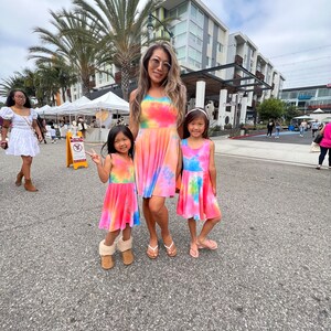 May include: Three people wearing tie-dye dresses in pink, blue, and yellow colors. The woman is wearing a long dress, while the two girls are wearing shorter dresses. They are standing on a paved street in front of a building.