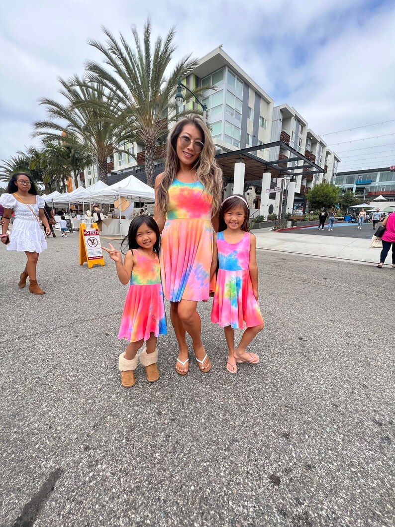 May include: Three people wearing tie-dye dresses in pink, blue, and yellow colors. The woman is wearing a long dress, while the two girls are wearing shorter dresses. They are standing on a paved street in front of a building.