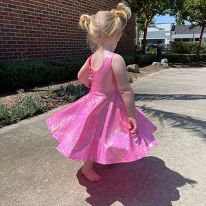 May include: A young girl wearing a pink iridescent dress with a flared skirt. She is standing on a concrete surface with a brick wall in the background.