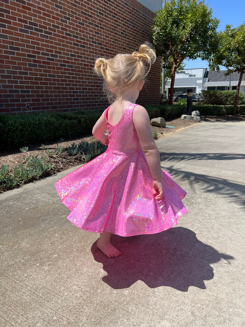 May include: A young girl wearing a pink iridescent dress with a flared skirt. She is standing on a concrete surface with a brick wall in the background.