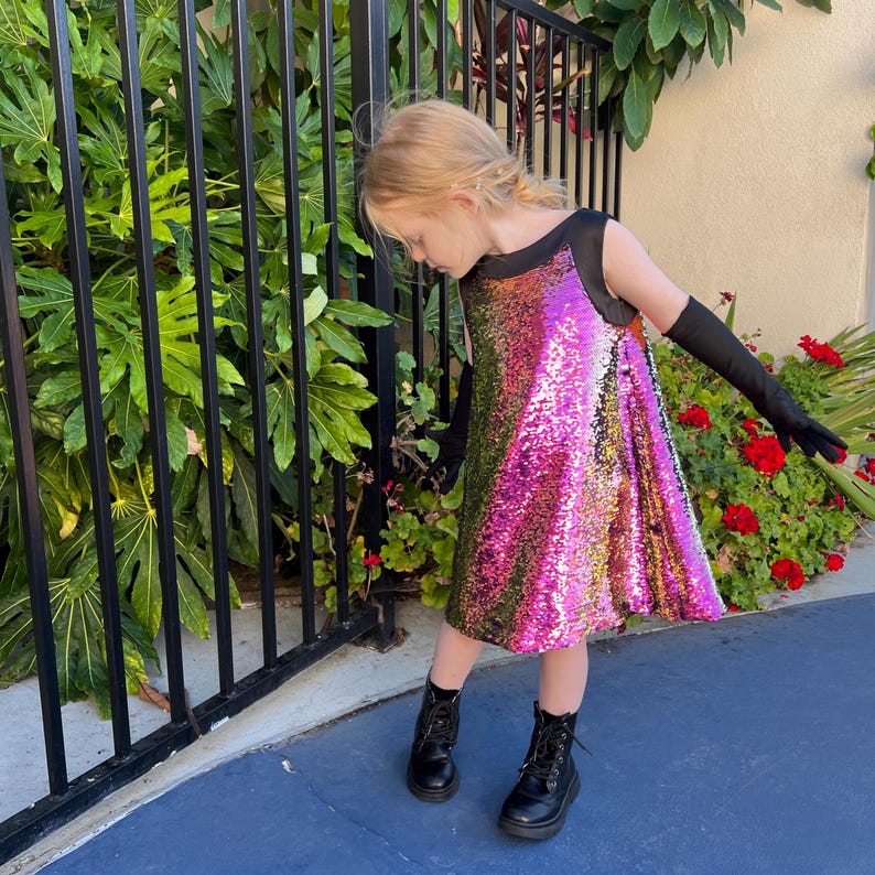 May include: A young person wearing a pink and green sequined dress with black sleeves and black boots. The dress has a black bodice and a flared skirt.