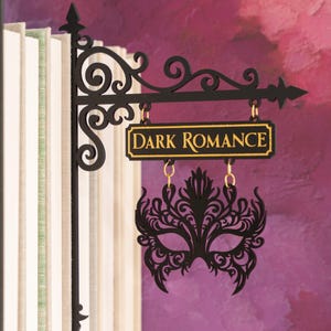 Può includere: Un'insegna in metallo nero con la scritta "Dark Romance" in lettere dorate, appesa a una staffa decorativa. Sotto l'insegna è appesa una maschera nera e ornata. L'insegna è accanto a una fila di libri.