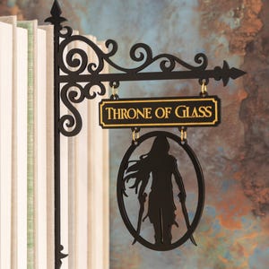 May include: Black metal sign with the text "THRONE OF GLASS" in gold lettering. The sign features a silhouette of a figure holding two swords within an oval frame. The sign is hanging from a decorative bracket.