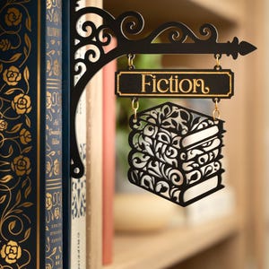 May include: A black metal bookshelf divider featuring a decorative scrollwork bracket that extends outward. It holds a hanging rectangular sign with the word Fiction in gold lettering above a stack of three laser-cut books.