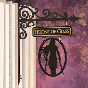 May include: Black metal sign with the text "THRONE OF GLASS" in gold lettering. The sign features a silhouette of a figure holding two swords within an oval frame, hanging from a decorative bracket.