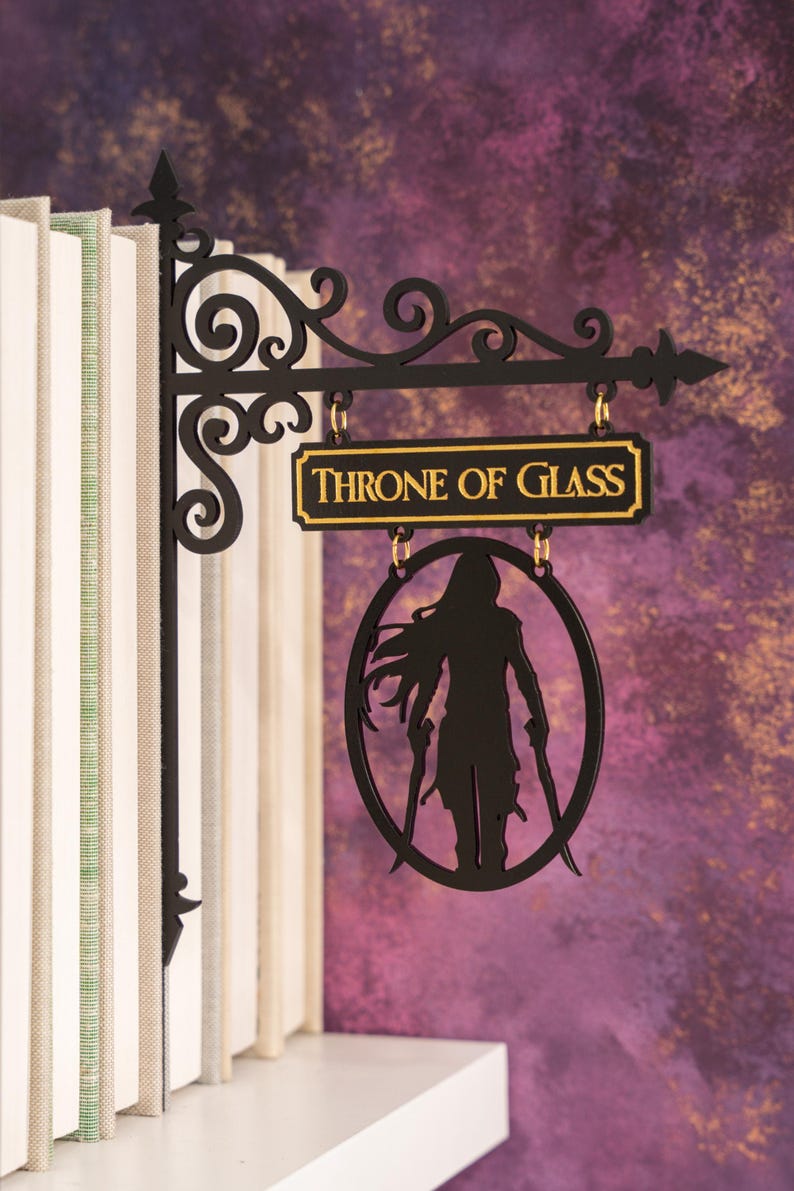 May include: Black metal sign with the text "THRONE OF GLASS" in gold lettering. The sign features a silhouette of a figure holding two swords within an oval frame, hanging from a decorative bracket.