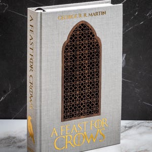 May include: A hardcover book titled "A Feast for Crows" by George R.R. Martin. The book has a light gray cover with gold lettering and a decorative wooden cutout. A gold bird emblem is on the spine.