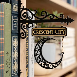 May include: Black metal sign with the words "CRESCENT CITY" in gold lettering. The sign features a crescent moon design and is hanging in front of a bookshelf with books.