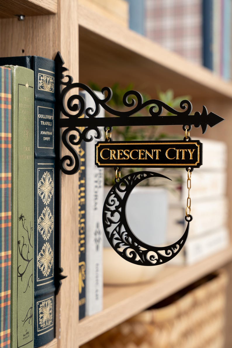 May include: Black metal sign with the words "CRESCENT CITY" in gold lettering. The sign features a crescent moon design and is hanging in front of a bookshelf with books.
