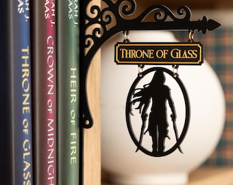 Könnte beinhalten: Ein dekorativer Buchständer aus schwarzem Metall mit einer verzierten Schnörkelhalterung, an der ein rechteckiges Schild mit dem Text Throne of Glass sowie ein runder Anhänger mit der Silhouette einer Figur mit zwei Schwertern hängt.