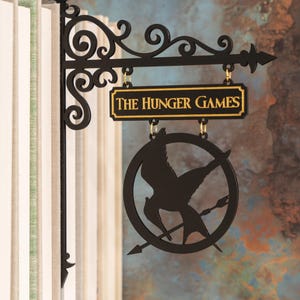 May include: Black metal sign with the text "THE HUNGER GAMES" in gold lettering. The sign features a circular emblem with a bird and arrow design, hanging from a decorative bracket.
