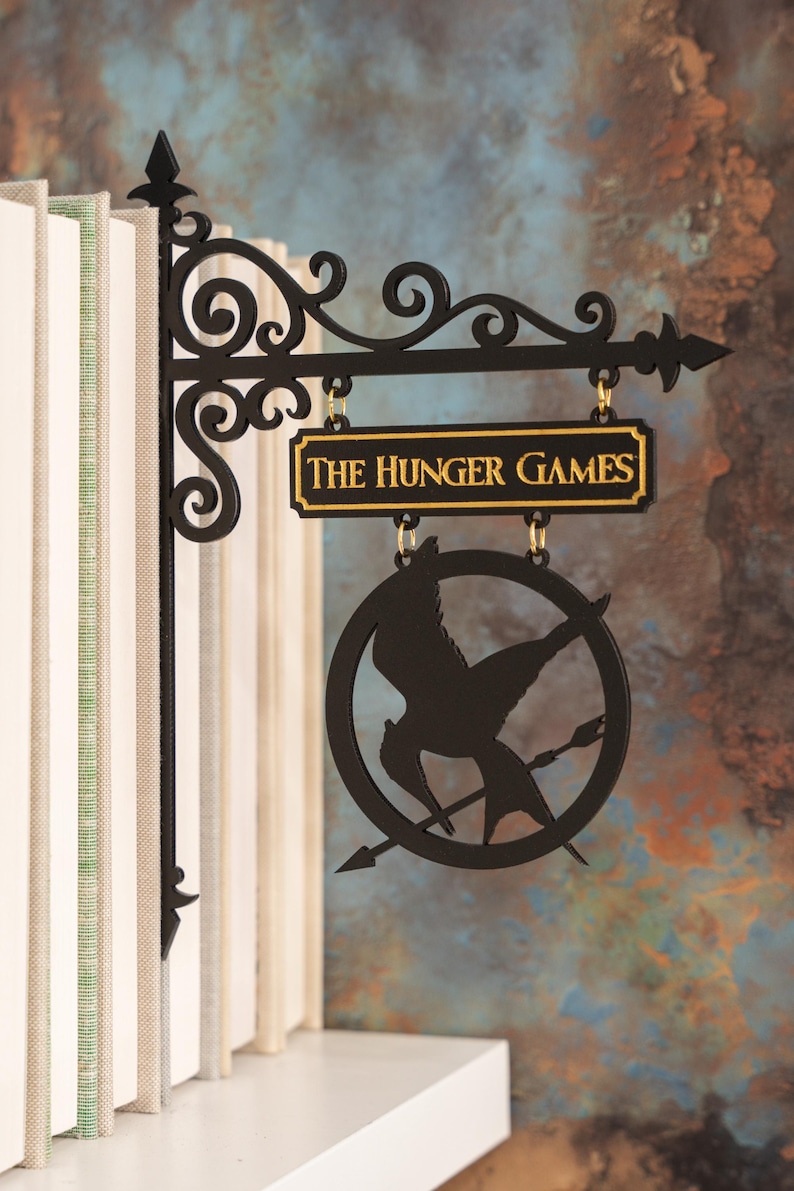 May include: Black metal sign with the text "THE HUNGER GAMES" in gold lettering. The sign features a circular emblem with a bird and arrow design, hanging from a decorative bracket.