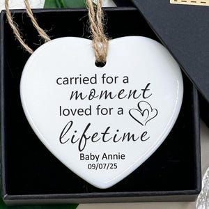 Pode incluir: Enfeite branco em forma de coração com o texto "carried for a moment, loved for a lifetime" e "Baby Annie 09/07/25". O enfeite está em uma caixa de presente preta com uma etiqueta "Hand made" e flores azuis.