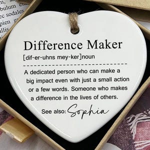 Może przedstawiać: Biała ceramiczna ozdoba w kształcie serca z tekstem "Difference Maker" i definicją słowa. Ozdoba zawiera również tekst "Zobacz także: Sophia".