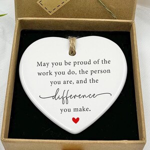 Könnte beinhalten: Weißer, herzförmiger Anhänger aus Keramik mit einem roten Herzen am Boden. Der Anhänger hat eine Schnur oben und trägt die Aufschrift "May you be proud of the work you do, the person you are, and the difference you make."