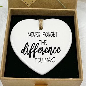 Puede incluir: Un adorno de cerámica blanca en forma de corazón con el texto "Nunca olvides la diferencia que haces" en letras negras. El adorno está en una caja de regalo marrón con una pequeña etiqueta que dice "Hecho a mano".