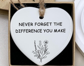 Op de afbeelding: Een wit hartvormig ornament met de tekst "VERGEET NOOIT HET VERSCHIL DAT JE MAAKT" en een bloemenillustratie. Het hangt aan een touwlus en wordt gepresenteerd in een geschenkdoos met een kleine bloemdecoratie.
