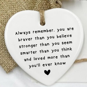 Puede incluir: Adorno de cerámica blanca en forma de corazón con una cuerda negra colgando de la parte superior. El adorno tiene una inscripción negra que dice "Recuerda siempre. Eres más valiente de lo que crees más fuerte de lo que pareces más inteligente de lo que piensas y amado más de lo que jamás sabrás".