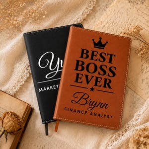 May include: Two rectangular notebooks with stitched edges. The black notebook features white text reading "YU" and "MARKETING". The tan notebook features a black crown icon, the text "BEST BOSS EVER", a star, the name "Brynn", and "FINANCE ANALYST".