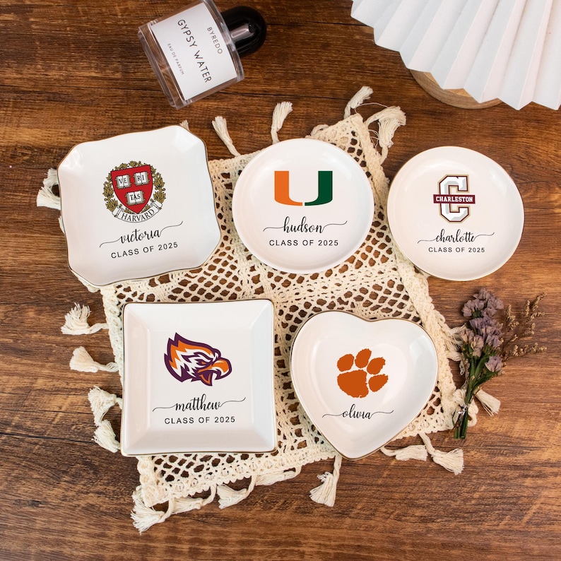 May include: A collection of white ceramic trinket dishes in various shapes, including square, round, and heart. Each dish features a college logo and a name with "CLASS OF 2025". Gold trim accents the edges. A perfume bottle is in the background.