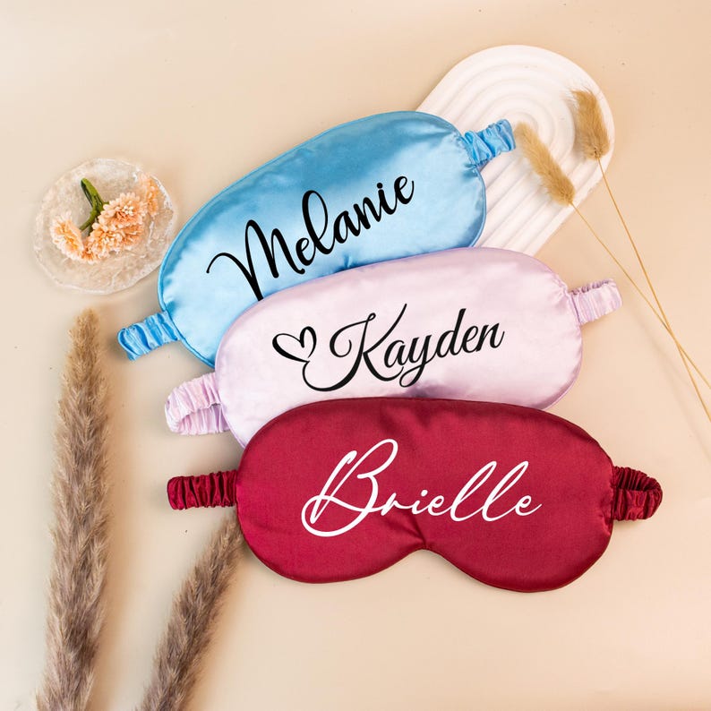 May include: Three personalized satin sleep masks in blue, pink, and burgundy. Each mask has a name written in cursive: "Melanie", "Kayden", and "Brielle". The masks are arranged on a neutral-toned surface.